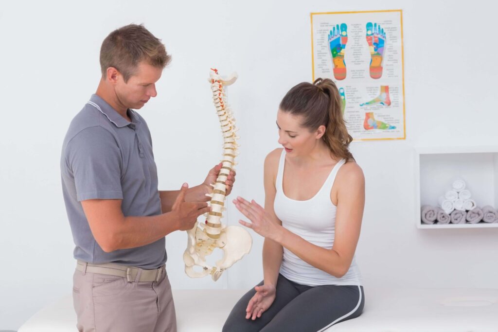 A male physiotherapist holding a model of the spine explains to a female patient sitting on the table what is affecting her back pain.