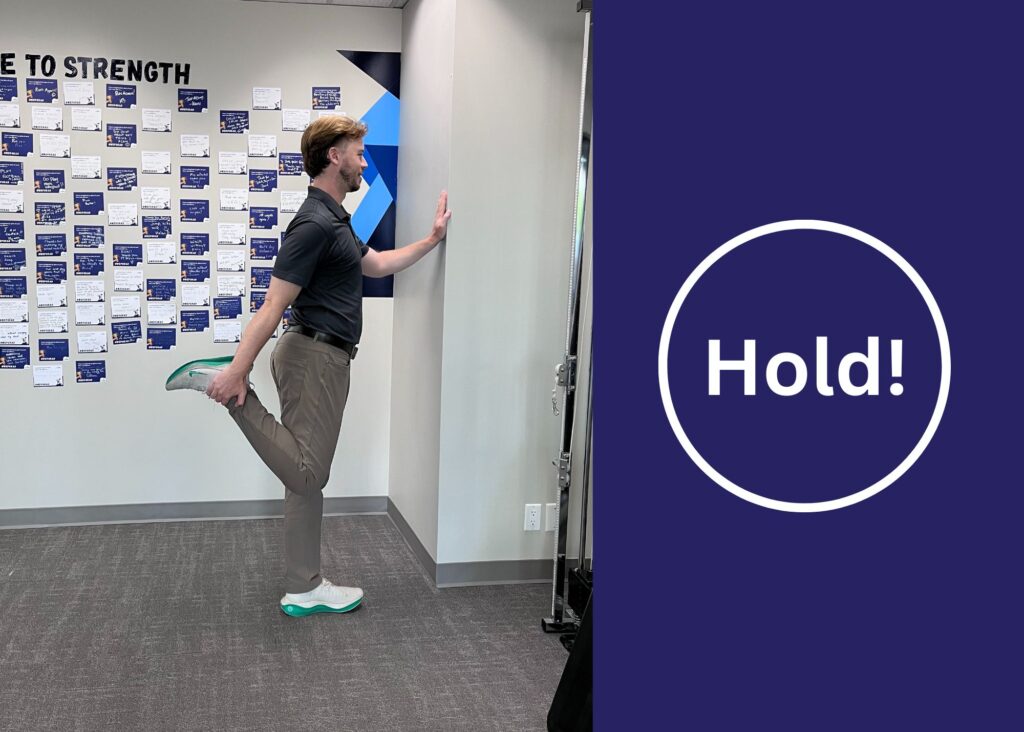 Chiropractor Dr. Donny demonstrating the quad stretch. He is standing and supporting himself on the wall while holding his foot behind him. There is also text on the image that says 'hold'