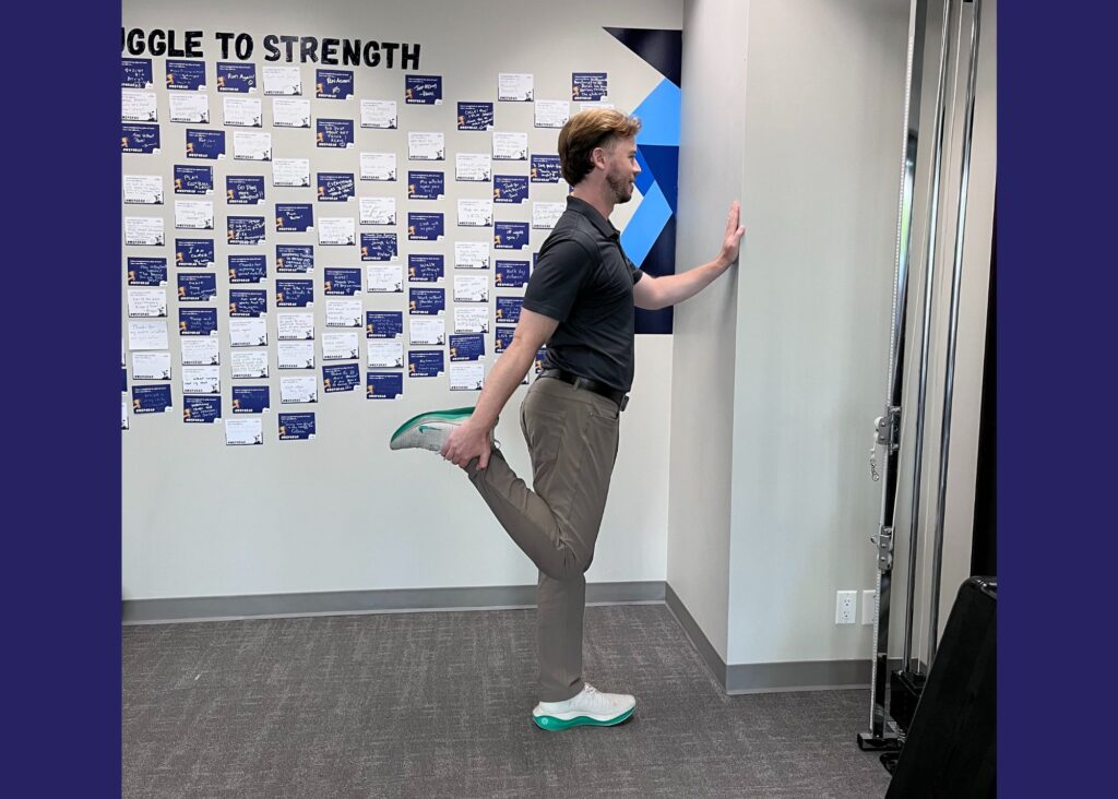 Chiropractor Dr. Donny demonstrating the quad stretch. He is standing and supporting himself on the wall while holding his foot behind him.