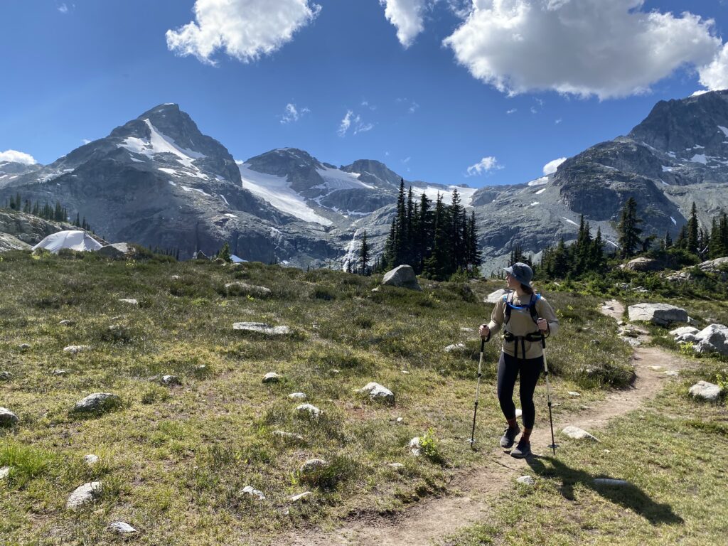 Hiker walking on a well defined trail wearing a small hiking pack and using trekking/hiking poles. The hiker is in front of an alpine scene with mountains, snow, trees and rocks.
