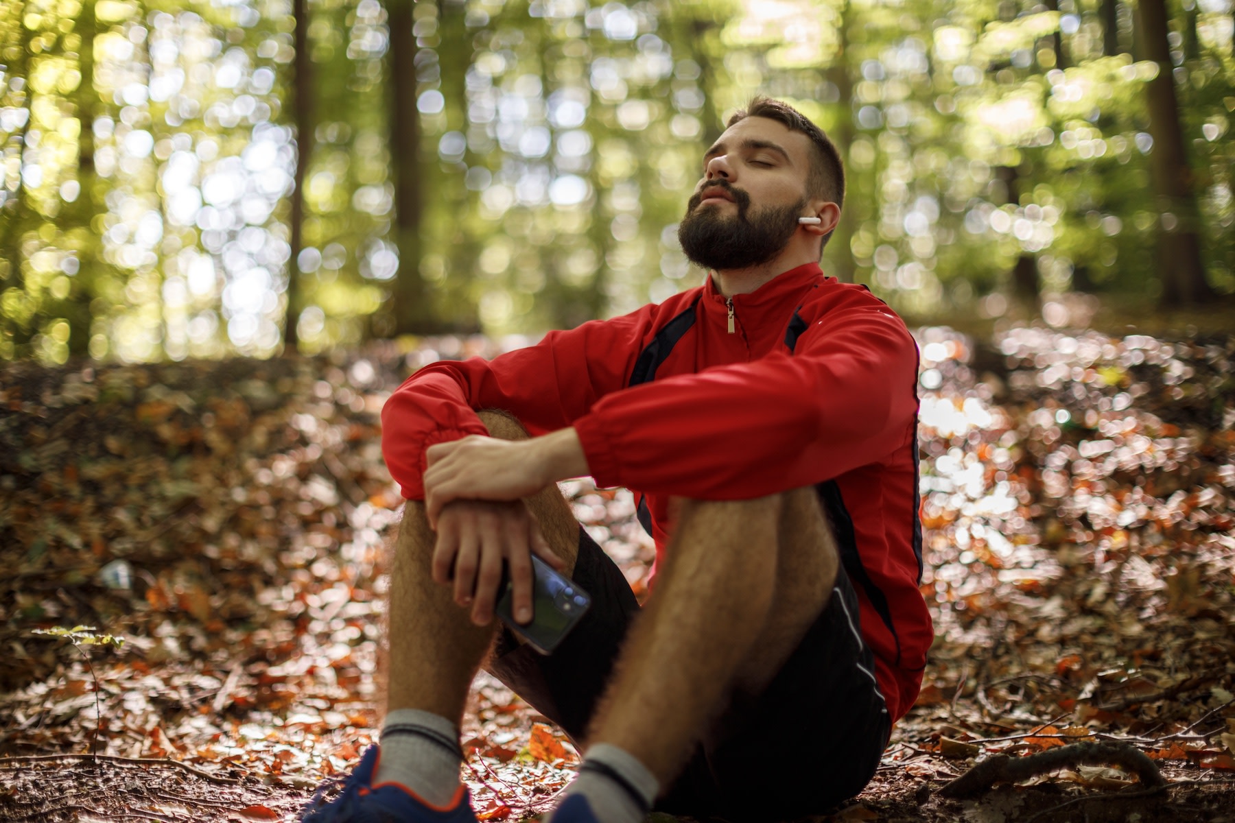 A man sitting in the woods with his face up towards the sky. His eyes are closed and he is in a meditative position. He is wearing a red running jacket, shorts and running shoes.